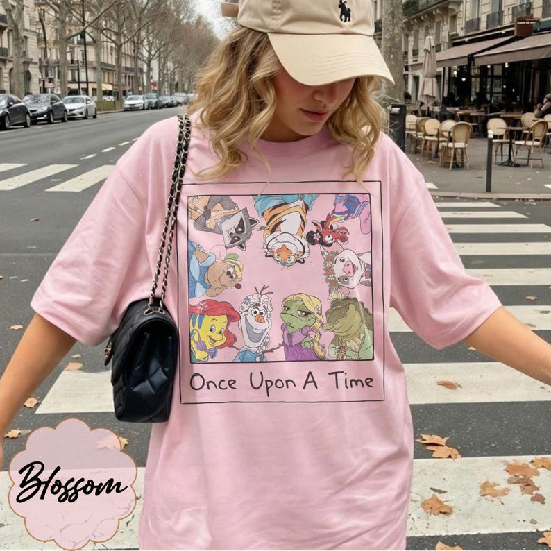 May include: A light pink t-shirt featuring a framed graphic of cartoon characters with the text "Once Upon A Time." The shirt is worn with a beige cap and a black purse with a chain strap. The word "Blossom" is written in cursive at the bottom left.