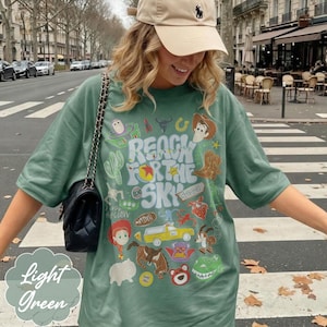 Pode incluir: Camiseta verde claro com o texto "Reach for the Sky" e gr&aacute;ficos de personagens de desenhos animados. A camiseta tem gola redonda e mangas curtas. Uma bolsa preta com al&ccedil;a de corrente &eacute; vis&iacute;vel.