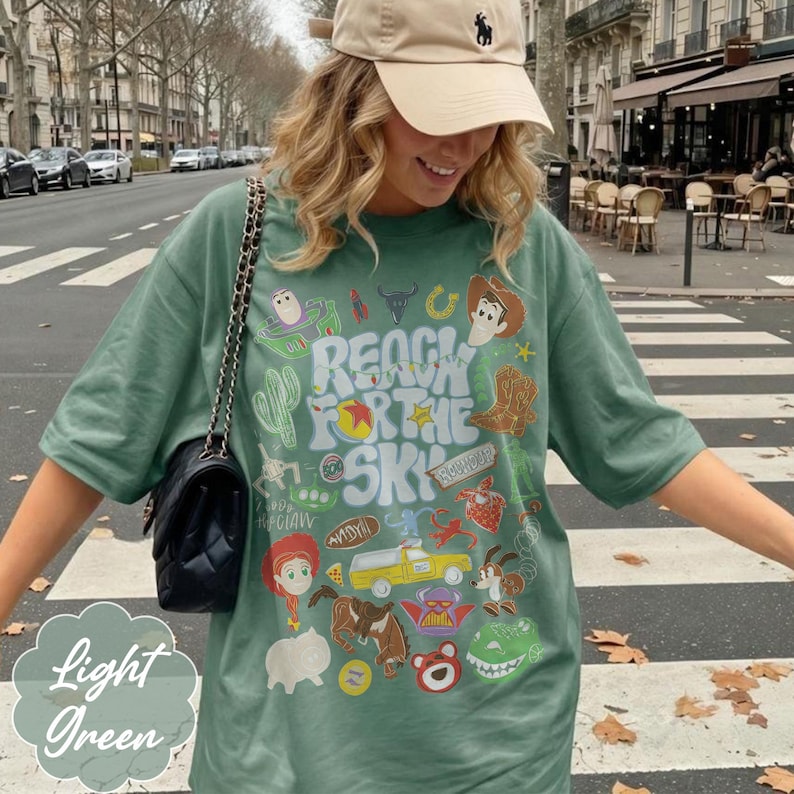 Pode incluir: Camiseta verde claro com o texto "Reach for the Sky" e gr&aacute;ficos de personagens de desenhos animados. A camiseta tem gola redonda e mangas curtas. Uma bolsa preta com al&ccedil;a de corrente &eacute; vis&iacute;vel.
