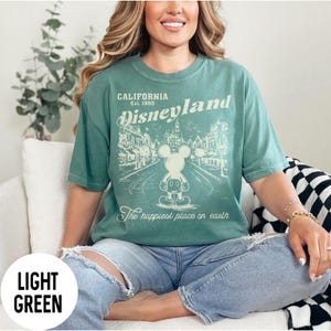 Może przedstawiać: Jasnozielona koszulka z grafiką Disneyland. Grafika zawiera tekst "California Est. 1955 Disneyland" oraz rysunek Myszki Miki przed sceną z parku. Zawiera r&oacute;wnież tekst "The happiest place on earth".