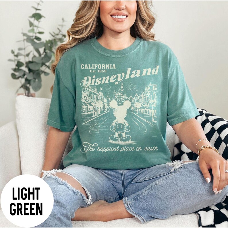 Może przedstawiać: Jasnozielona koszulka z grafiką Disneyland. Grafika zawiera tekst "California Est. 1955 Disneyland" oraz rysunek Myszki Miki przed sceną z parku. Zawiera r&oacute;wnież tekst "The happiest place on earth".