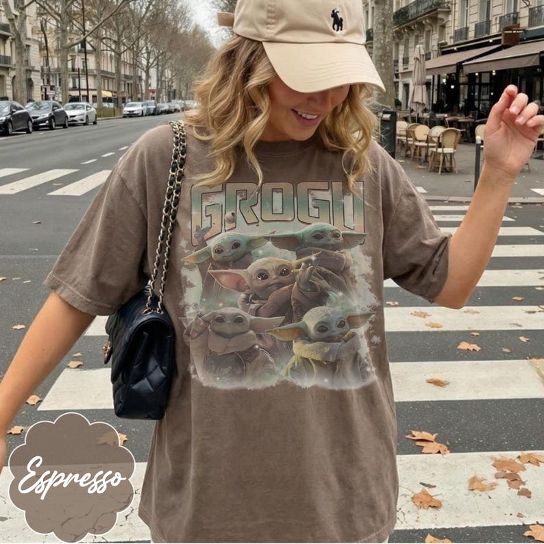 May include: A brown short-sleeve t-shirt featuring a graphic print of multiple Grogu characters and the text "GROGU" in a vintage style. The shirt has a crew neckline and a relaxed fit, paired with a black quilted shoulder bag with a chain strap.