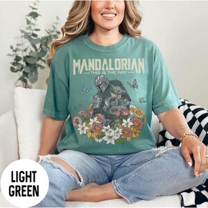 Puede incluir: Camiseta verde claro con el texto "MANDALORIAN - THIS IS THE WAY" sobre un gr&aacute;fico del Mandaloriano y Grogu rodeados de flores y mariposas. La camiseta es de color verde claro.