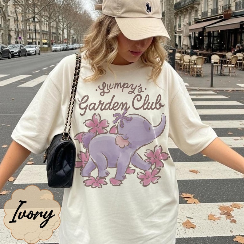 Op de afbeelding: Ivoor t-shirt met een paarse olifant afbeelding omringd door roze bloemen en de tekst "Jumpy's Garden Club". Het shirt wordt gedragen met een beige baseball cap.