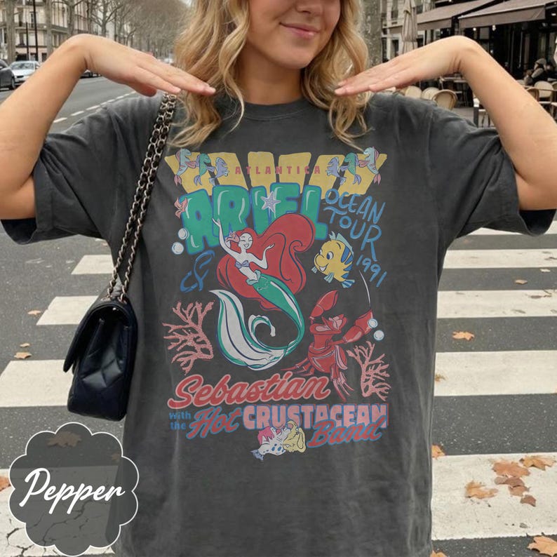 May include: Dark gray t-shirt featuring a colorful graphic of Ariel the mermaid, Flounder, and Sebastian from The Little Mermaid. The shirt has the text "Atlantica Ocean Tour 1991" and "Sebastian with the Hot Crustacean Band".