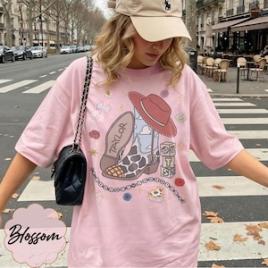 May include: A light pink short-sleeve t-shirt featuring a graphic print of a cowboy boot with a cow print pattern, a red hat, and blocks with the letters T, S, and the number 13. The text Taylor is on the boot sole, surrounded by small floral icons.