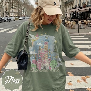 Puede incluir: Camiseta verde musgo con un gr&aacute;fico de castillo colorido, un hada y el texto "Happiest Birthday on Earth". La camiseta tiene mangas cortas y un ajuste relajado. Se ve un bolso negro con una correa de cadena.