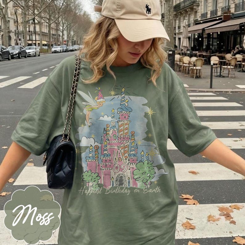 Puede incluir: Camiseta verde musgo con un gr&aacute;fico de castillo colorido, un hada y el texto "Happiest Birthday on Earth". La camiseta tiene mangas cortas y un ajuste relajado. Se ve un bolso negro con una correa de cadena.