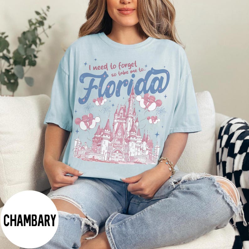 May include: Light blue t-shirt with the text "I need to forget so take me to Florida" in blue script. The shirt features a pink and white illustration of a castle and Mickey Mouse balloons. The shirt is worn by a person.
