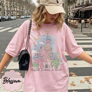 Puede incluir: Camiseta rosa claro con una ilustraci&oacute;n colorida de un castillo, un hada y el texto "Happiest Birthday on Earth". La camiseta tiene mangas cortas y un corte holgado.