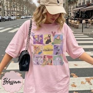 May include: A light pink short-sleeve t-shirt featuring a grid of colorful illustrations of characters from the movie The Emperor's New Groove, including the film title text in the center, paired with a black quilted shoulder bag with a chain strap.