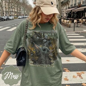 Puede incluir: Una camiseta verde oliva con un estampado gr&aacute;fico de un personaje mandaloriano y una peque&ntilde;a criatura verde. El texto dice THE MANDALORIAN y THIS IS THE WAY. Se lleva un bolso de hombro negro acolchado con una correa de cadena dorada.