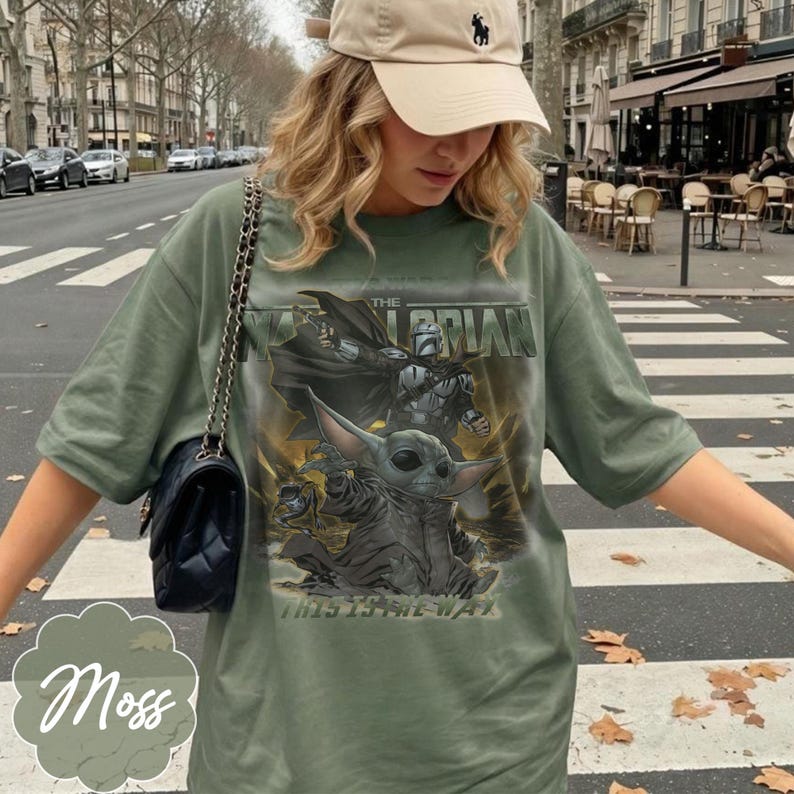Puede incluir: Una camiseta verde oliva con un estampado gr&aacute;fico de un personaje mandaloriano y una peque&ntilde;a criatura verde. El texto dice THE MANDALORIAN y THIS IS THE WAY. Se lleva un bolso de hombro negro acolchado con una correa de cadena dorada.