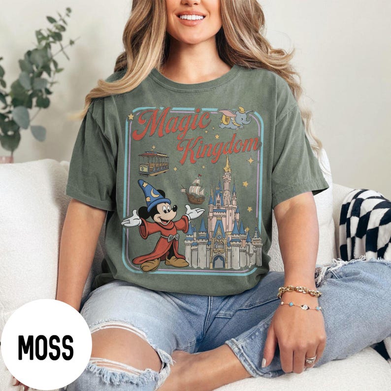 May include: Moss green t-shirt with a retro "Magic Kingdom" graphic. The design includes Mickey Mouse in a sorcerer's outfit, a castle, and other theme park elements. The shirt has a relaxed fit and is made of a soft material.