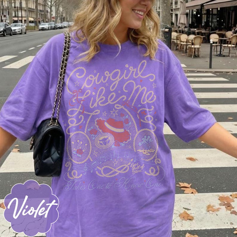 May include: A violet short-sleeved t-shirt featuring a graphic print in light yellow and red. The design includes the text "Cowgirl Like Me", "Eyes full of stars...", and "Takes One to Know One", alongside illustrations of a hat, rope, and musical notes.