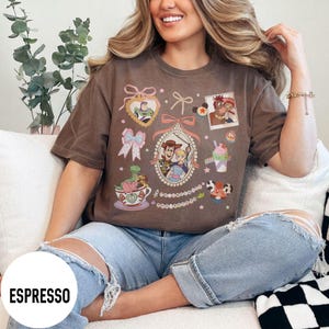 May include: Espresso-colored t-shirt featuring cartoon characters and decorative elements. The design includes a central oval frame with Woody and Bo Peep, and other characters like Buzz Lightyear and Rex. The shirt has a relaxed fit.