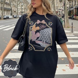 May include: Black t-shirt featuring a colorful graphic design with a bow, fireworks, and a geometric sphere. The graphic also includes a cartoon character. The shirt has short sleeves and a relaxed fit.