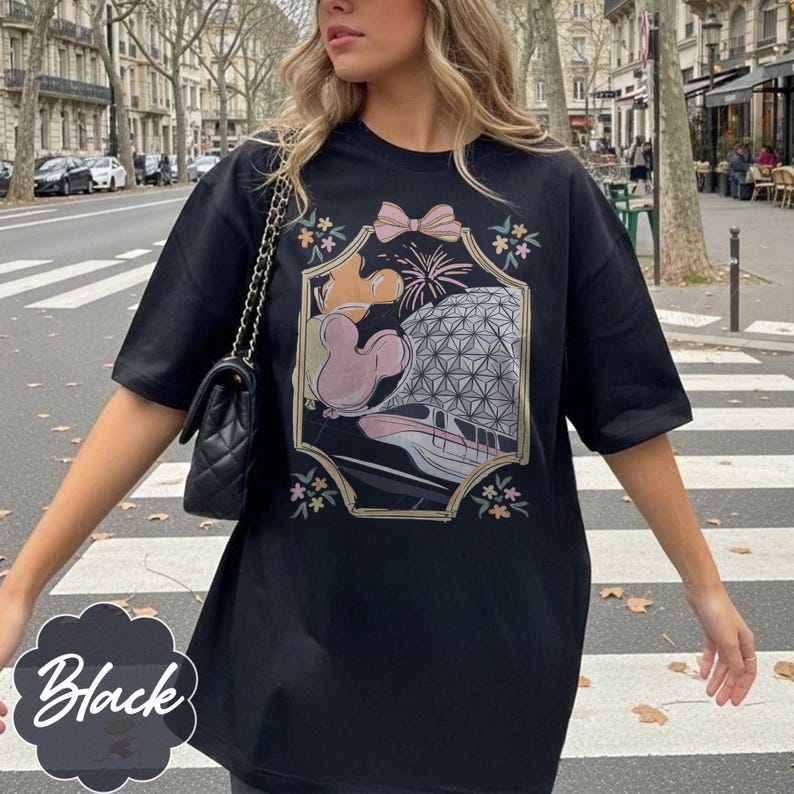 May include: Black t-shirt featuring a colorful graphic design with a bow, fireworks, and a geometric sphere. The graphic also includes a cartoon character. The shirt has short sleeves and a relaxed fit.