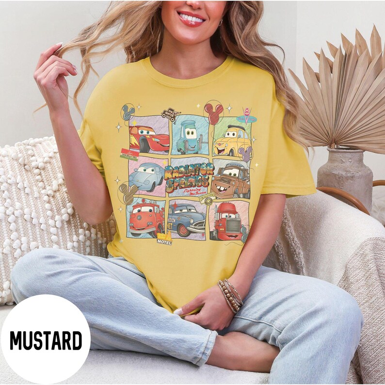 May include: Mustard-colored t-shirt featuring a grid of cartoon cars from the movie "Cars." The design includes Lightning McQueen and other characters, with the text "Radiator Springs" and "Lightning McQueen." The shirt is a relaxed fit.