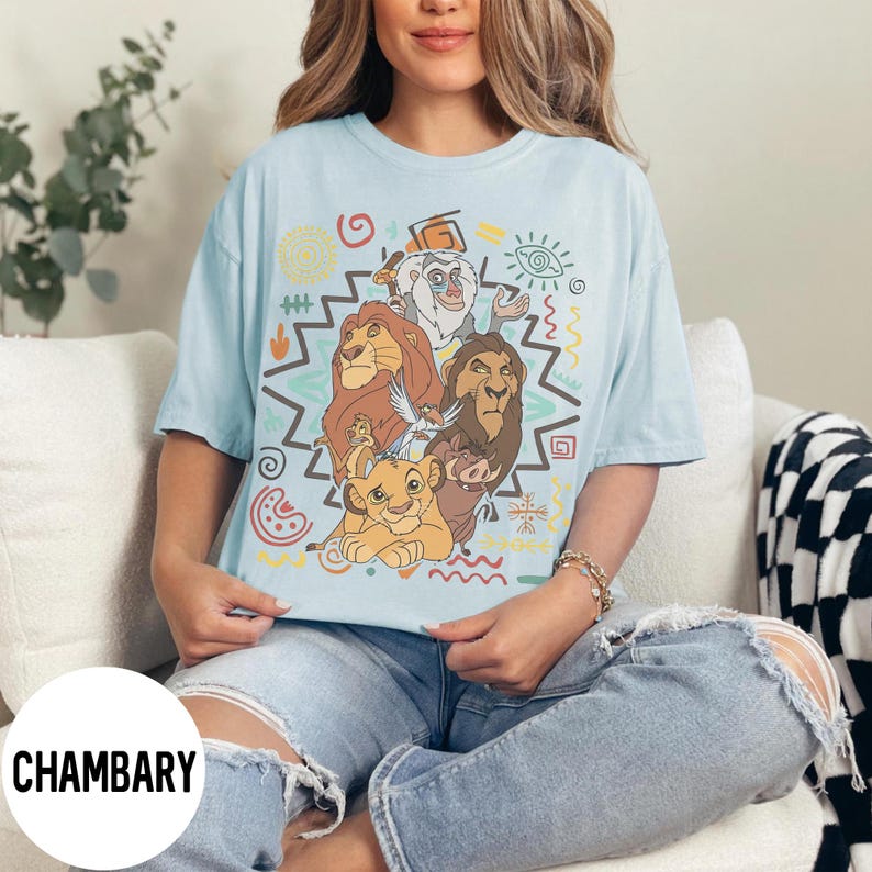 Puede incluir: Camiseta azul claro con un dise&ntilde;o de personajes de El Rey Le&oacute;n, incluyendo a Simba y Mufasa, y formas geom&eacute;tricas coloridas. La palabra "CHAMBARY" est&aacute; impresa en la parte inferior izquierda.