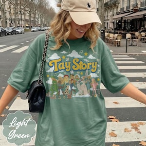 May include: A sage green short-sleeve t-shirt featuring a graphic print with the yellow text Tay Story. The design includes a series of illustrated figures in various outfits, clouds, stars, and musical notes. A black quilted shoulder bag is present.