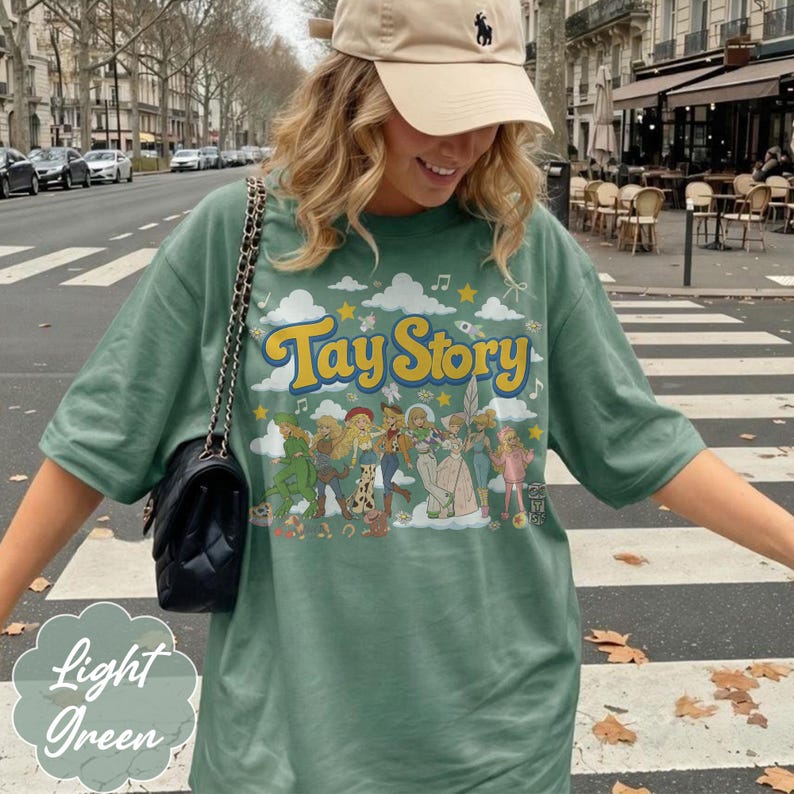 May include: A sage green short-sleeve t-shirt featuring a graphic print with the yellow text Tay Story. The design includes a series of illustrated figures in various outfits, clouds, stars, and musical notes. A black quilted shoulder bag is present.