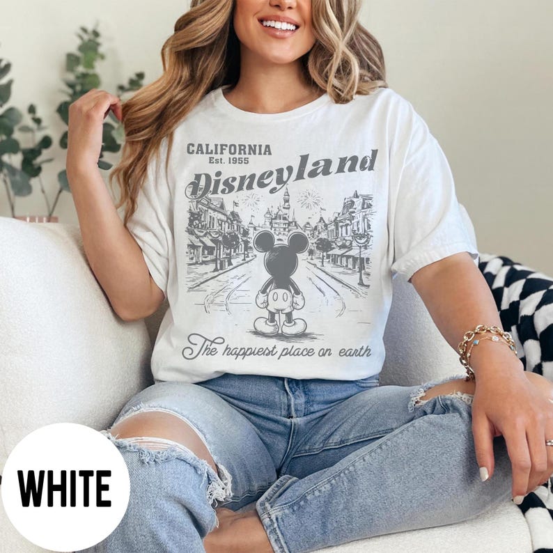 Może przedstawiać: Biała koszulka z grafiką Disneyland. Projekt przedstawia Myszka Miki, tekst "CALIFORNIA Est. 1955 Disneyland" i "The happiest place on earth". Koszulka ma luźny kr&oacute;j.