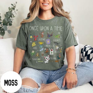 May include: A moss green t-shirt with the words "ONCE UPON A TIME" and cartoon illustrations of Disney characters. The shirt features characters like Ariel, Jasmine, and Cinderella. The word "MOSS" is printed on the bottom left.