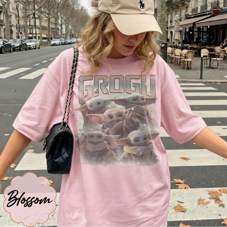 May include: A light pink t-shirt featuring a graphic print of multiple Grogu characters and the text "GROGU" in a vintage-style font. The shirt has short sleeves and a crew neckline, paired with a black quilted shoulder bag with a chain strap.