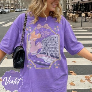 May include: A violet-colored t-shirt with a graphic design featuring a bow, fireworks, Mickey Mouse balloons, and a monorail. The word "Violet" is printed in a cursive font at the bottom left of the shirt.