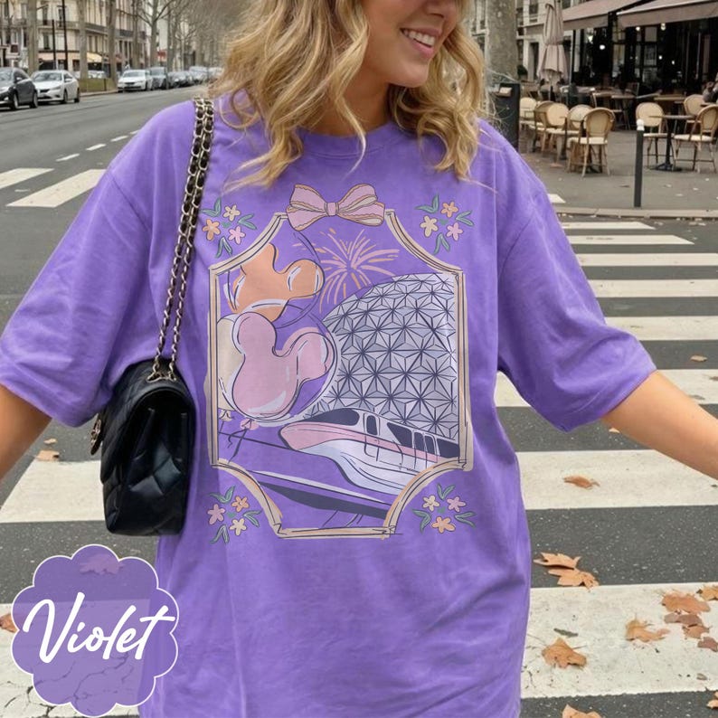 May include: A violet-colored t-shirt with a graphic design featuring a bow, fireworks, Mickey Mouse balloons, and a monorail. The word "Violet" is printed in a cursive font at the bottom left of the shirt.
