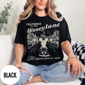 Może przedstawiać: Czarny t-shirt z grafiką Disneyland. Projekt zawiera tekst "CALIFORNIA Est. 1955 Disneyland" i "The happiest place on earth" z ilustracją Myszki Miki. Koszulka jest noszona przez osobę.