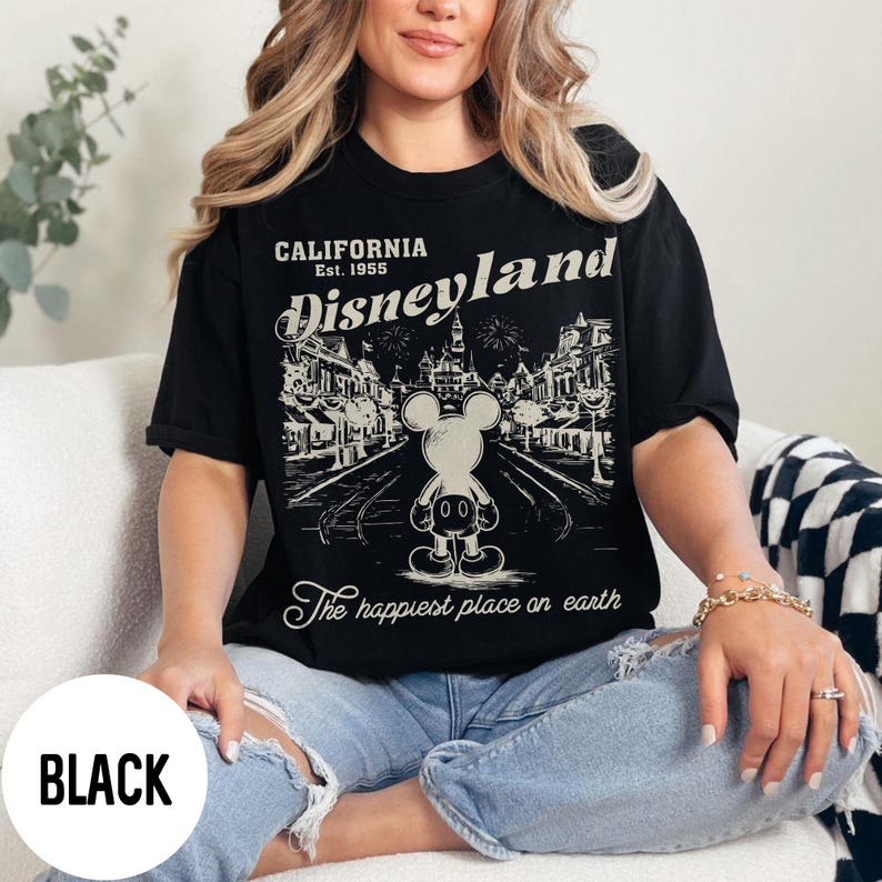 Może przedstawiać: Czarny t-shirt z grafiką Disneyland. Projekt zawiera tekst "CALIFORNIA Est. 1955 Disneyland" i "The happiest place on earth" z ilustracją Myszki Miki. Koszulka jest noszona przez osobę.
