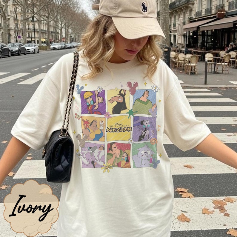 May include: An ivory short-sleeved t-shirt featuring a grid of colorful cartoon characters from The Emperor's New Groove, including Kuzco, Pacha, Yzma, and Kronk, with the movie title text in the center and small floral and icon accents.