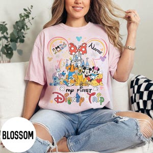 May include: A light pink t-shirt featuring a colorful graphic of Disney characters, a castle, and the text "my FIRST Disney TRIP." The design includes Mickey and Minnie Mouse, Goofy, Donald Duck, and other characters. The word "NAME" is also visible.