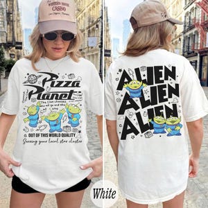 K&ouml;nnte beinhalten: Wei&szlig;es T-Shirt mit einem Pizza Planet-Motiv auf der Vorderseite und Alien-Charakteren auf der R&uuml;ckseite. Das Frontdesign enth&auml;lt den Text "Pizza Planet" und Cartoon-Aliens. Auf der R&uuml;ckseite steht das Wort "ALIEN" wiederholt.