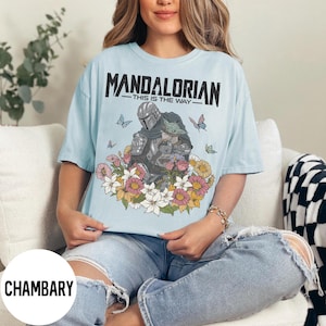 Puede incluir: Camiseta azul claro con un dise&ntilde;o floral con The Mandalorian y Grogu. El texto "MANDALORIAN - THIS IS THE WAY" est&aacute; impreso en la parte superior. Mariposas y flores rodean a los personajes.