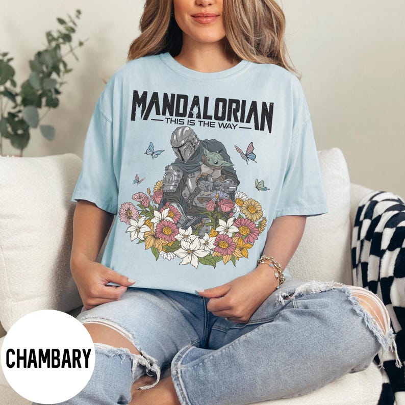 Puede incluir: Camiseta azul claro con un dise&ntilde;o floral con The Mandalorian y Grogu. El texto "MANDALORIAN - THIS IS THE WAY" est&aacute; impreso en la parte superior. Mariposas y flores rodean a los personajes.