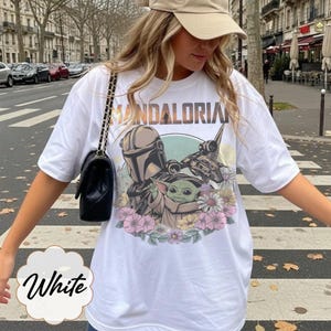 May include: White t-shirt featuring a graphic of The Mandalorian and Grogu surrounded by flowers. The word "MANDALORIAN" is printed across the top. The word "White" is written in cursive at the bottom of the shirt.