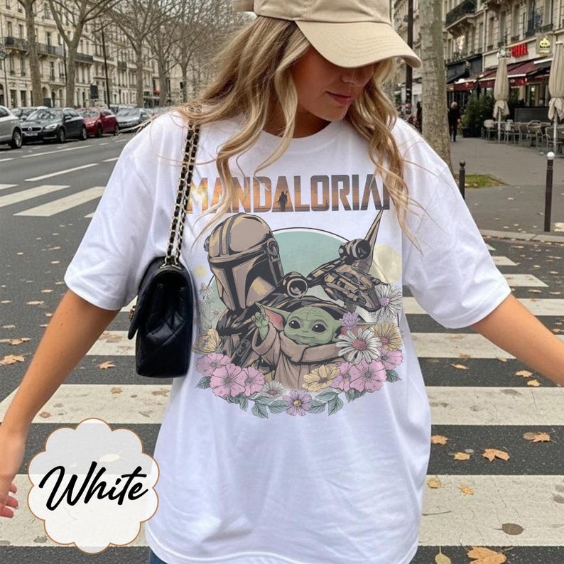 May include: White t-shirt featuring a graphic of The Mandalorian and Grogu surrounded by flowers. The word "MANDALORIAN" is printed across the top. The word "White" is written in cursive at the bottom of the shirt.