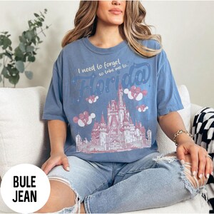 May include: A blue t-shirt with the text "I need to forget so take me to Florida" above a pink and white illustration of a castle. The shirt is worn with light blue jeans.