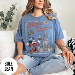 May include: A blue t-shirt with a graphic of Mickey Mouse as a wizard and the words "Magic Kingdom". The graphic includes a castle, a trolley, a ship, and Dumbo the elephant. The shirt is worn by a person.