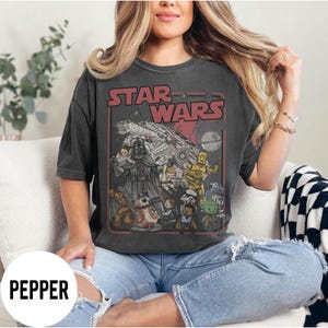 May include: Dark gray Star Wars t-shirt featuring a graphic of Darth Vader, the Millennium Falcon, and other characters. The text "STAR WARS" is in red letters. The shirt is a crew neck and has short sleeves.
