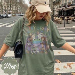 May include: An olive green short-sleeve t-shirt featuring a colourful graphic print of Toy Story characters including Woody, Buzz Lightyear, and a central green frog. The text Toy Story is printed in multicoloured letters above the character illustration.