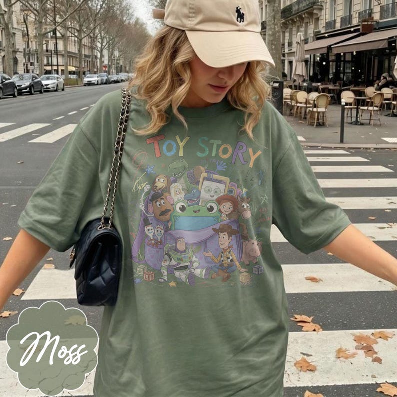 May include: An olive green short-sleeve t-shirt featuring a colourful graphic print of Toy Story characters including Woody, Buzz Lightyear, and a central green frog. The text Toy Story is printed in multicoloured letters above the character illustration.
