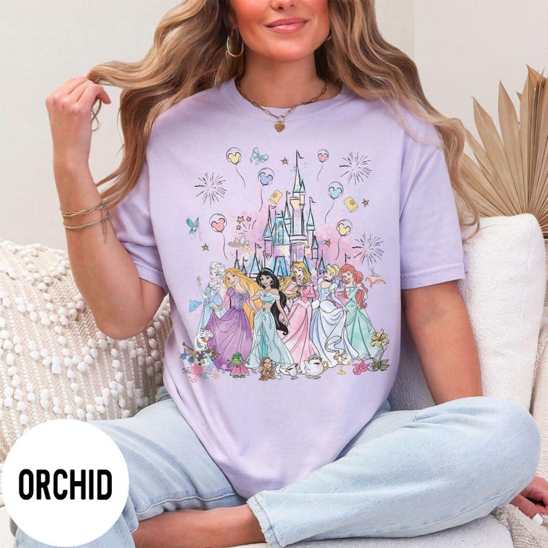 May include: A light purple t-shirt featuring a colorful illustration of a castle and several princesses. The word "ORCHID" is printed in a white circle at the bottom. The princesses are wearing gowns in various colors, and the castle is in the background.