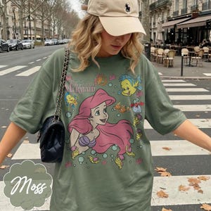 May include: Moss green t-shirt with a graphic of Ariel from Disney's The Little Mermaid. The design includes Ariel with pink hair, a purple seashell bra, and various sea creatures. The shirt has short sleeves and a relaxed fit.