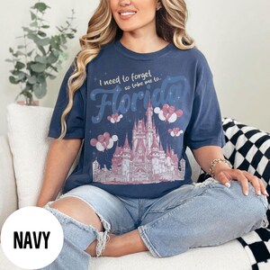 May include: Navy blue t-shirt with a graphic of a castle, balloons, and the word "Florida". The text "I need to forget so take me to..." is above the word "Florida". The shirt is a relaxed fit.