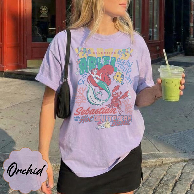 May include: A light purple t-shirt featuring a graphic of Ariel from The Little Mermaid, with the text "Ariel Ocean Tour 1991" and "Sebastian with the Hot Crustacean Band". The shirt is a vintage style.