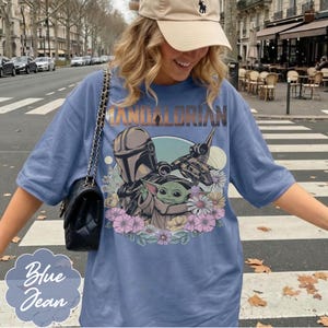 May include: A light blue t-shirt featuring a graphic of The Mandalorian and Grogu surrounded by flowers. The text "MANDALORIAN" is arched across the top. The shirt also has the text "Blue Jean" in a stylized font.